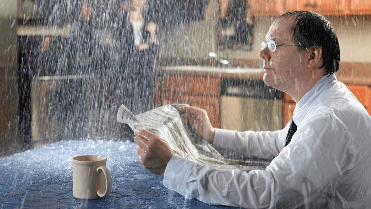 Man in a white shirt sitting at a table, shielding papers from heavy water spraying down indoors
