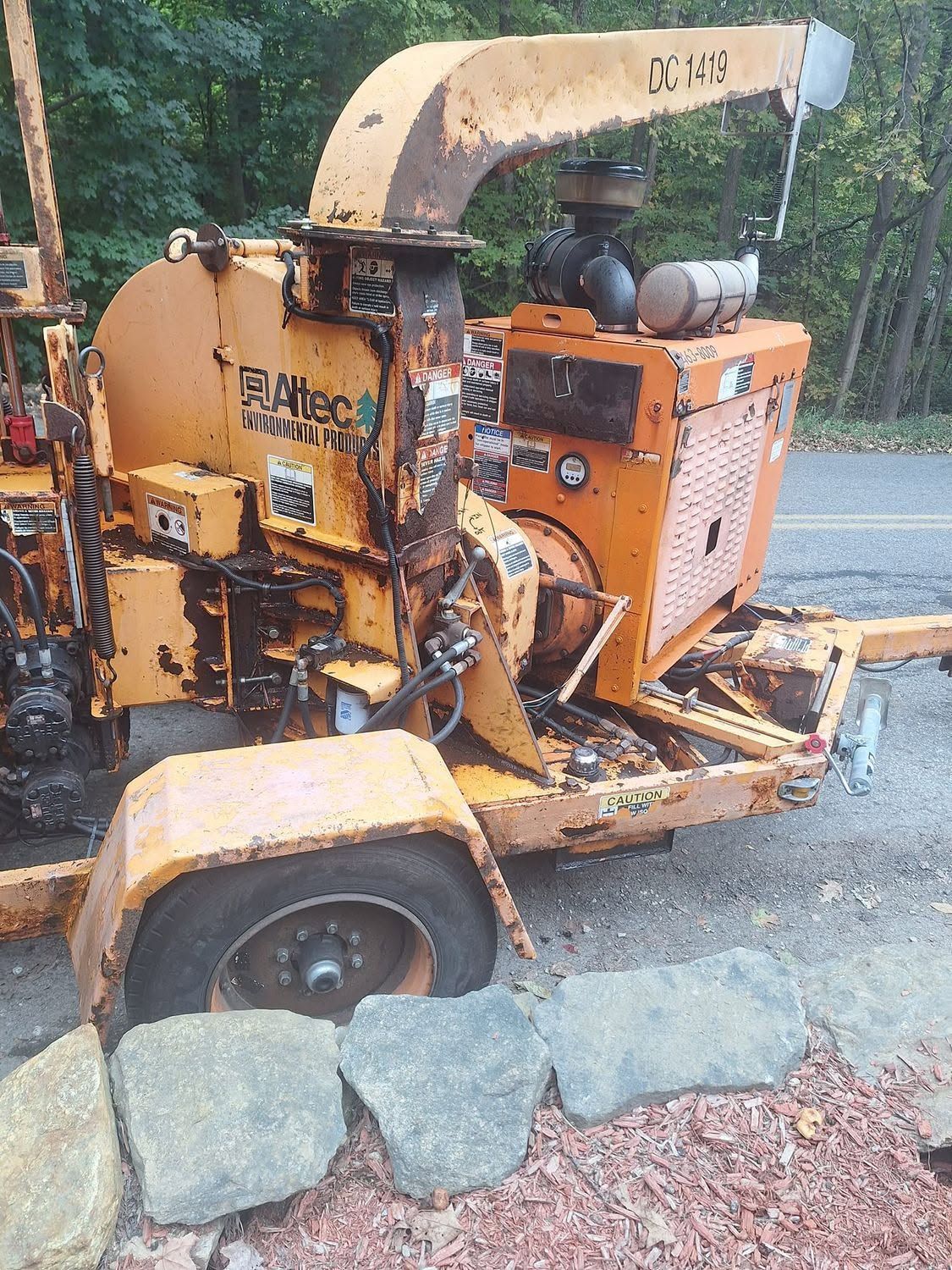 Orange wood chipper parked on a wet roadside beside stones and leaves.