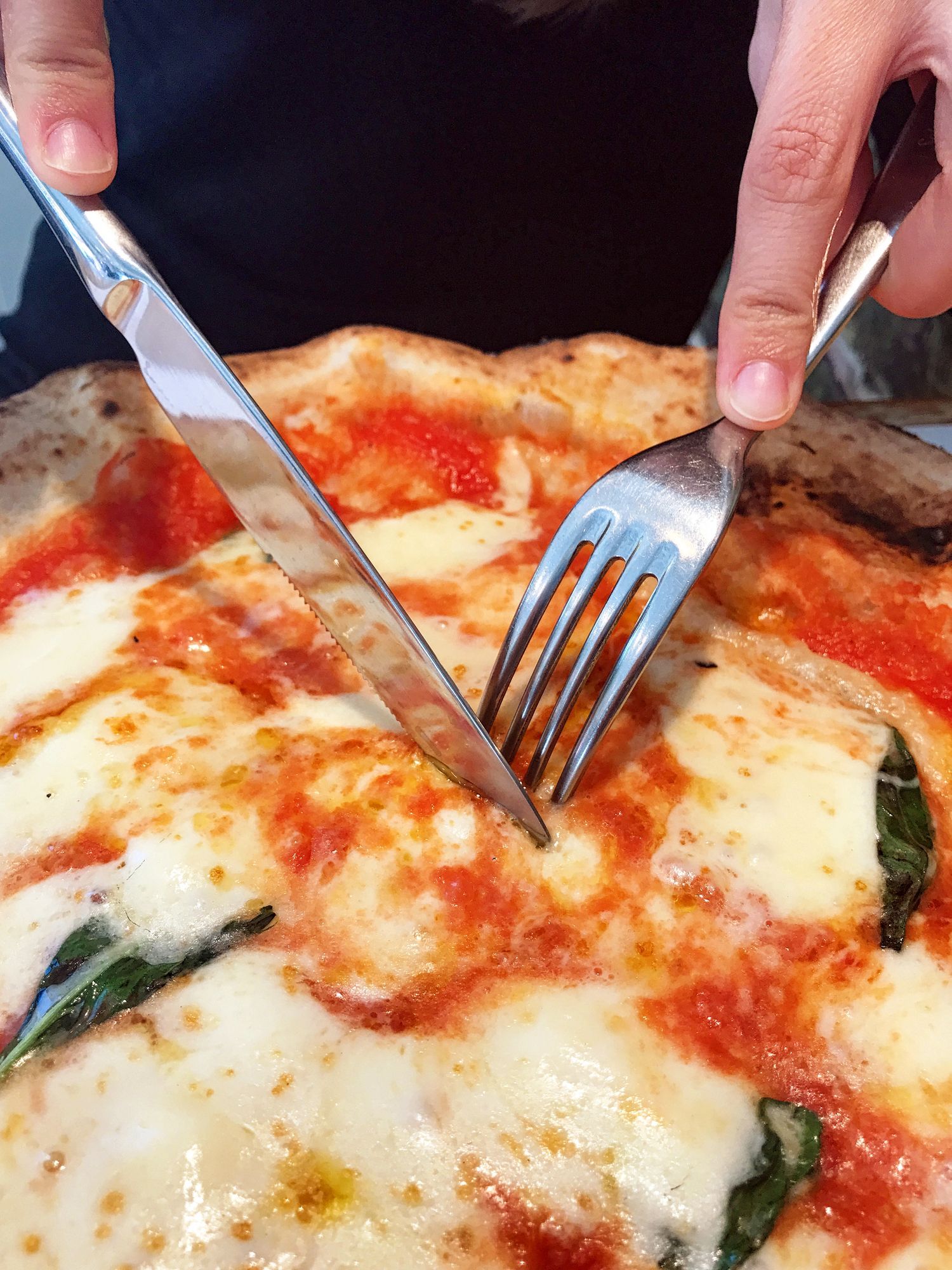 Persona che taglia una pizza con coltello e forchetta; primo piano.