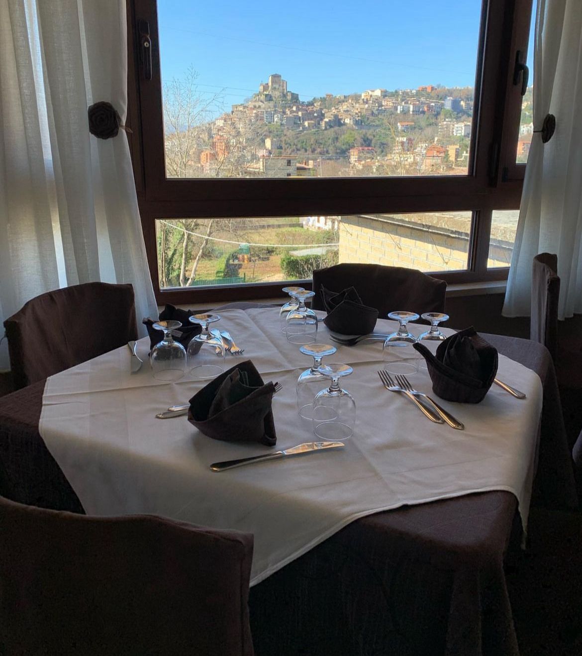Tavolo del ristorante apparecchiato con posate e bicchieri, con vista su una cittadina collinare.