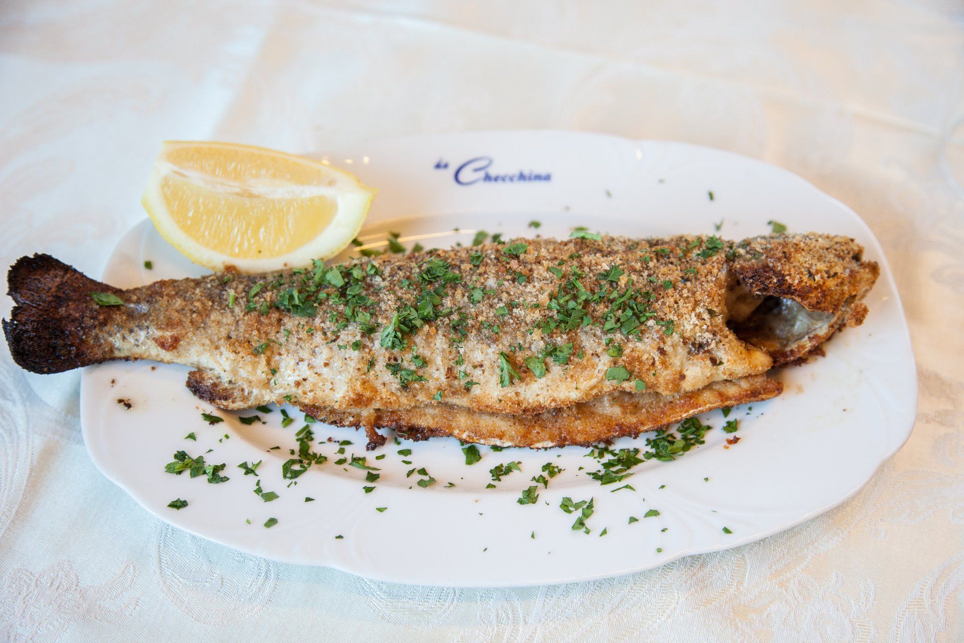 Pesce intero grigliato su un piatto bianco con guarnizione di fetta di limone e prezzemolo.
