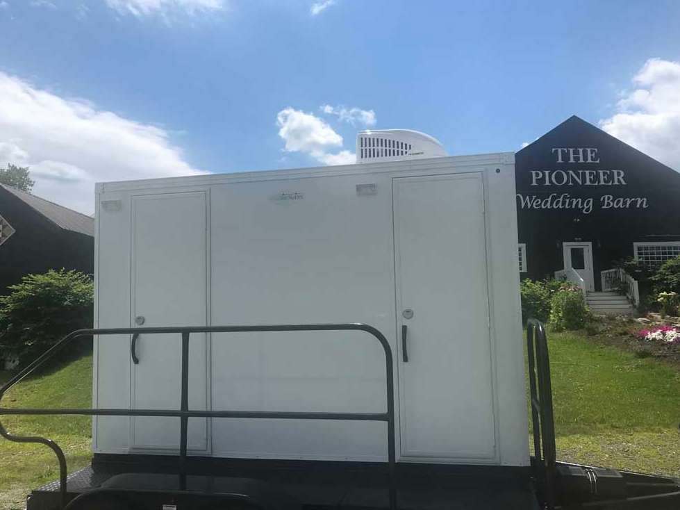 White portable restroom trailer on a trailer, with a barn in the background. Sunny day.