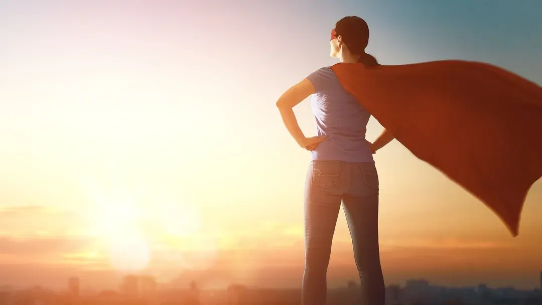 Une femme portant une cape de super-héros se tient devant une ville au coucher du soleil.
