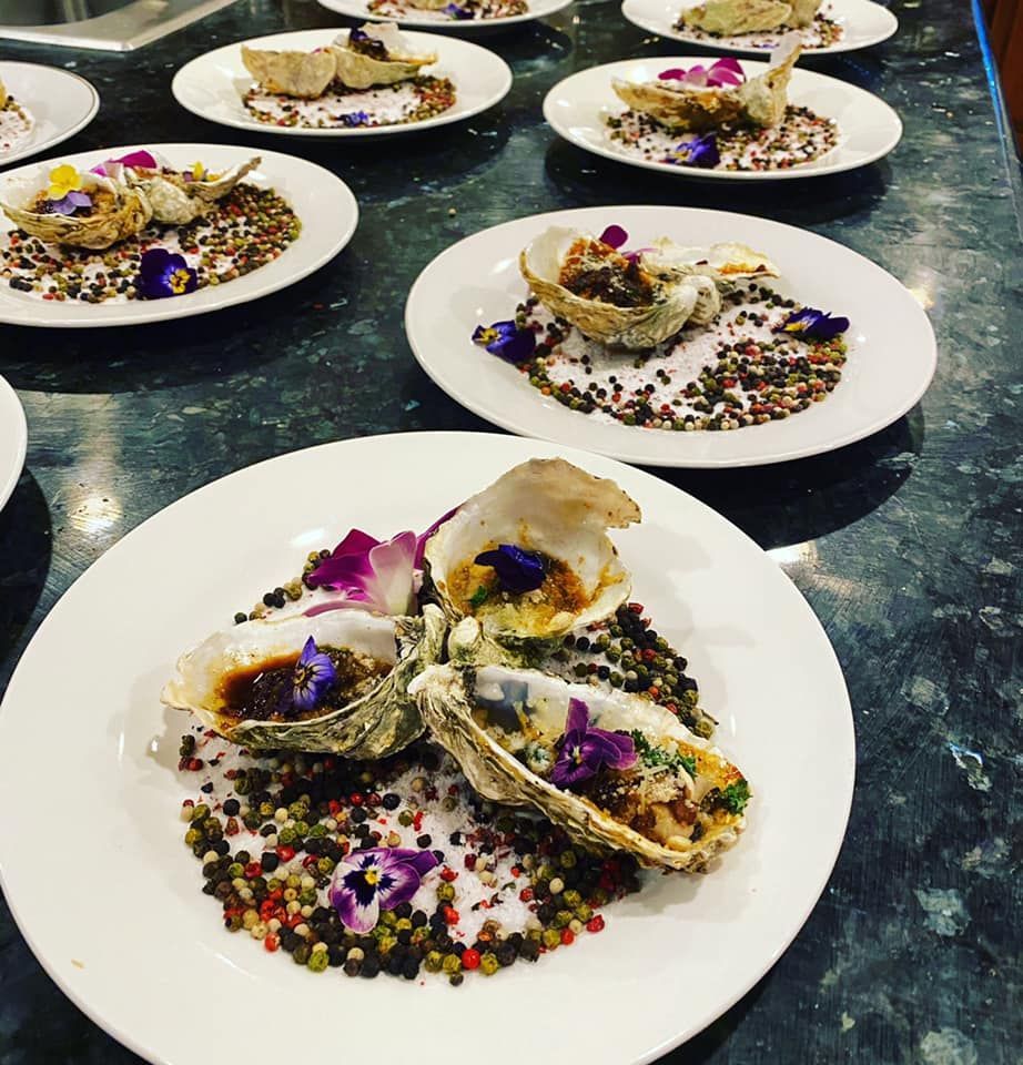 Plated oysters on a dark table, topped with edible flowers and colorful garnishes