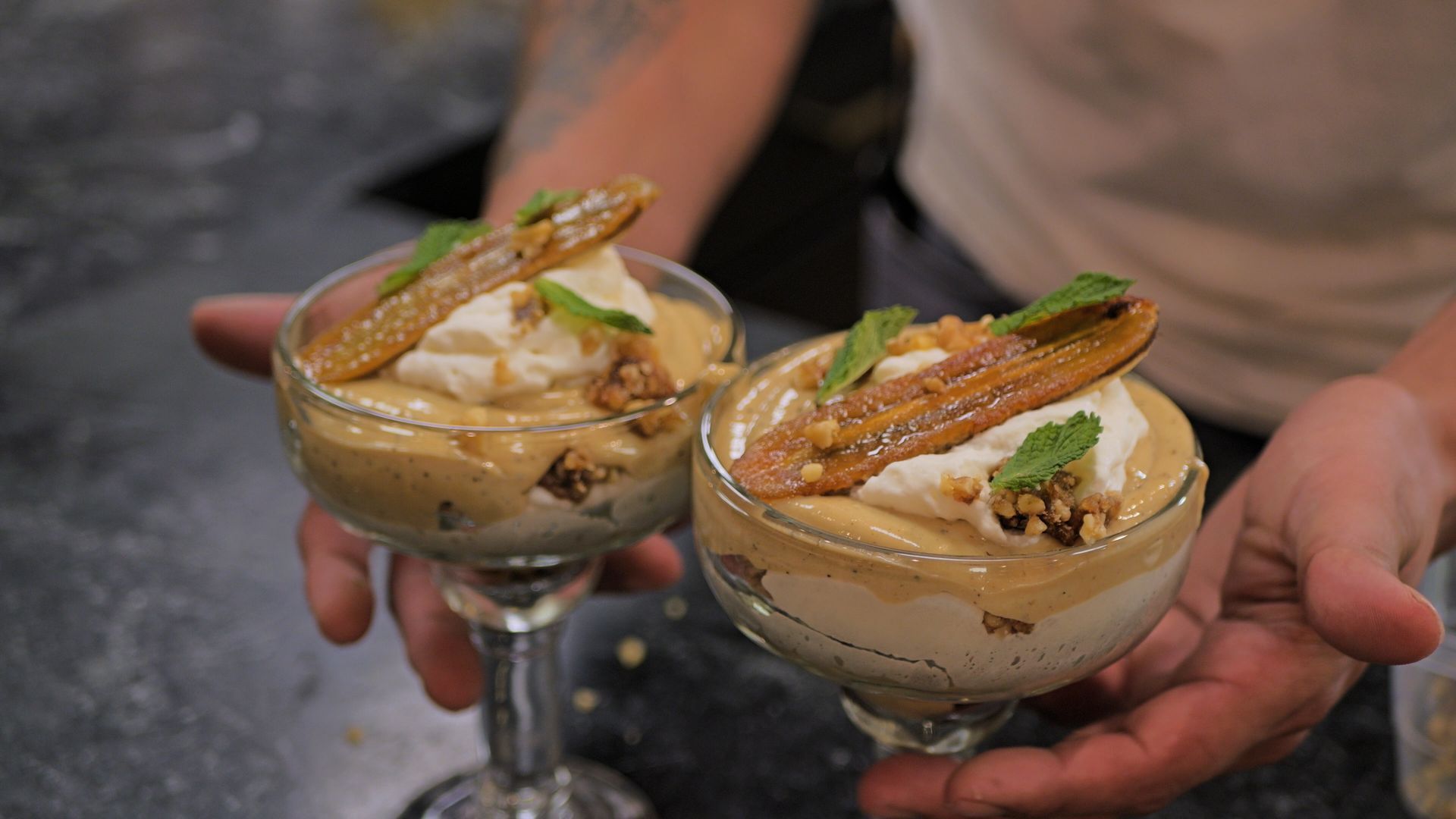Two dessert cocktails in coupe glasses, topped with mint and caramel garnish, held by a person’s hands.