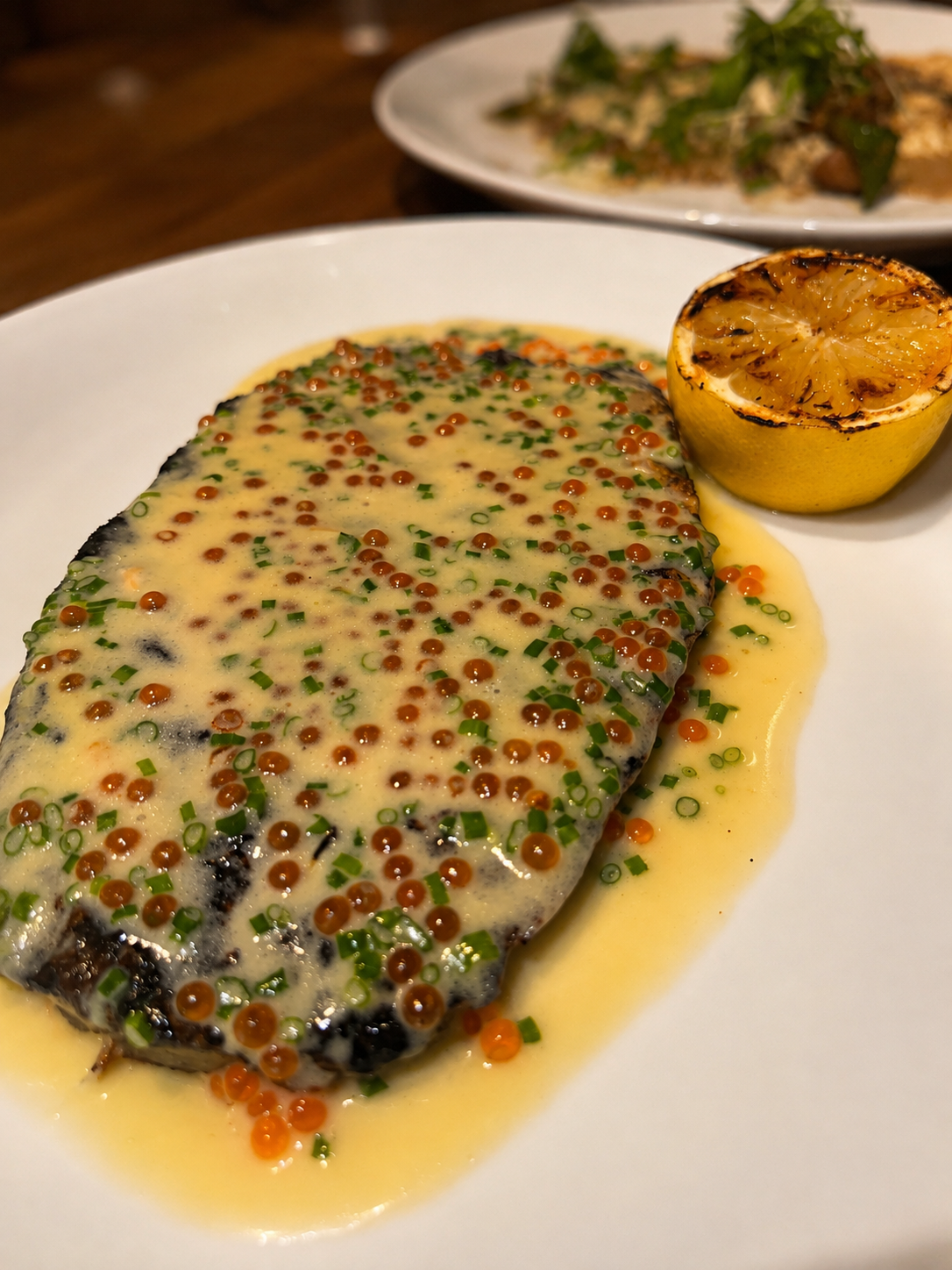 Fish fillet topped with creamy sauce and colorful caviar, served with a lemon half on a white plate