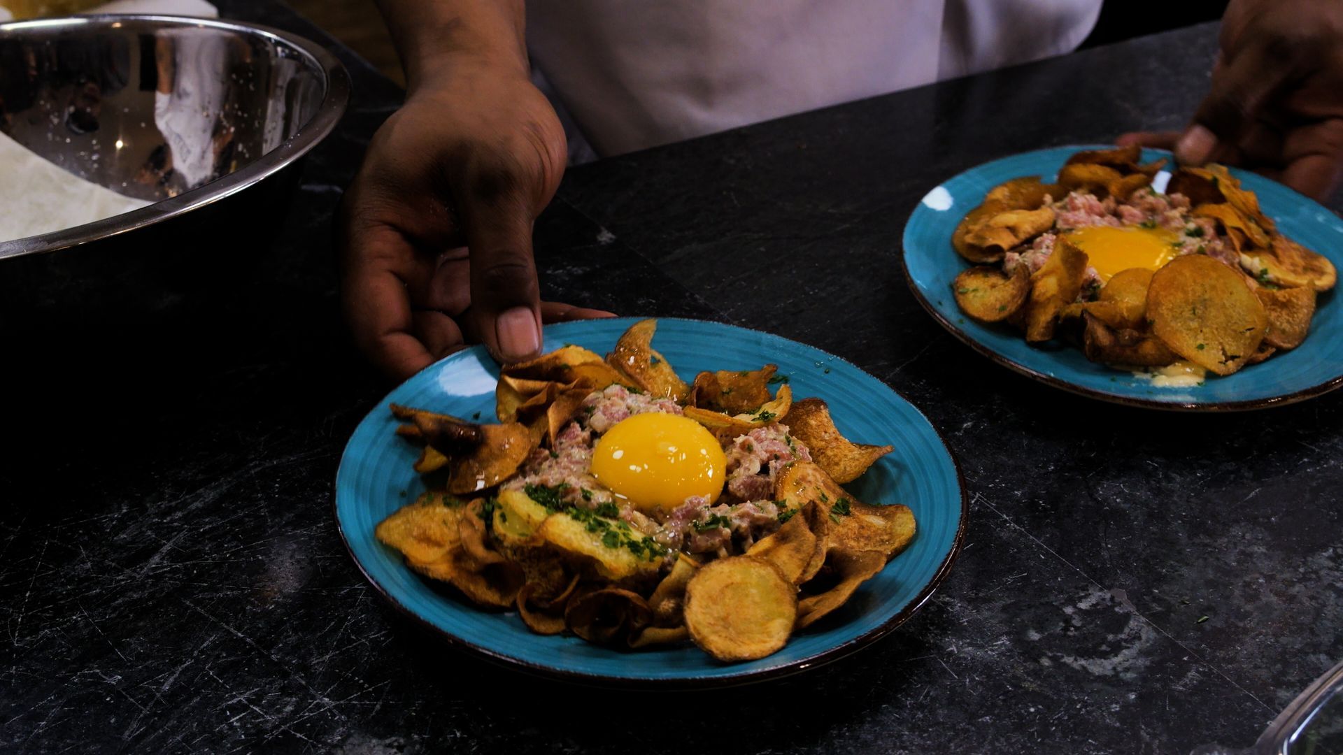 Two blue plates of fried food with sunny-side-up eggs on a dark counter, one being served by a hand