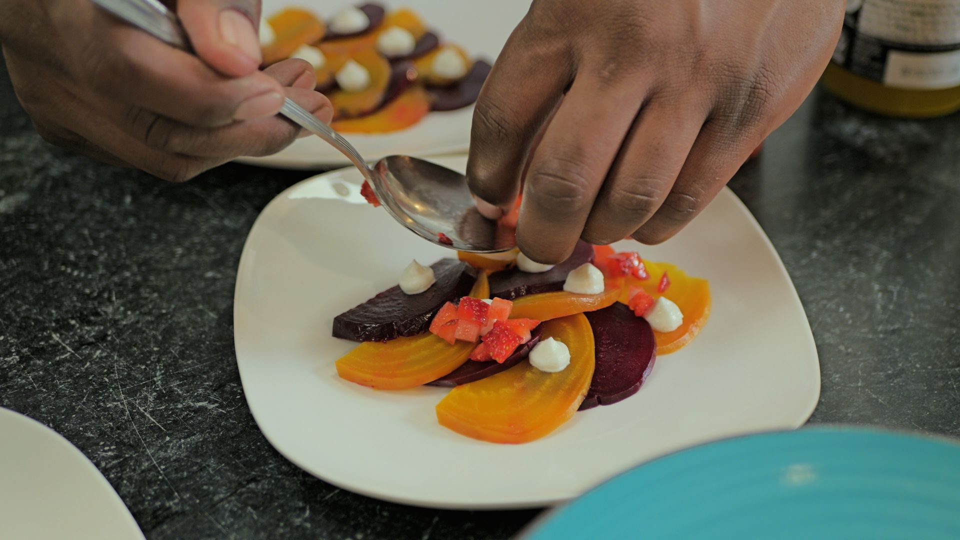 Hands arranging colorful sliced fruit and berries on a white plate with a spoon