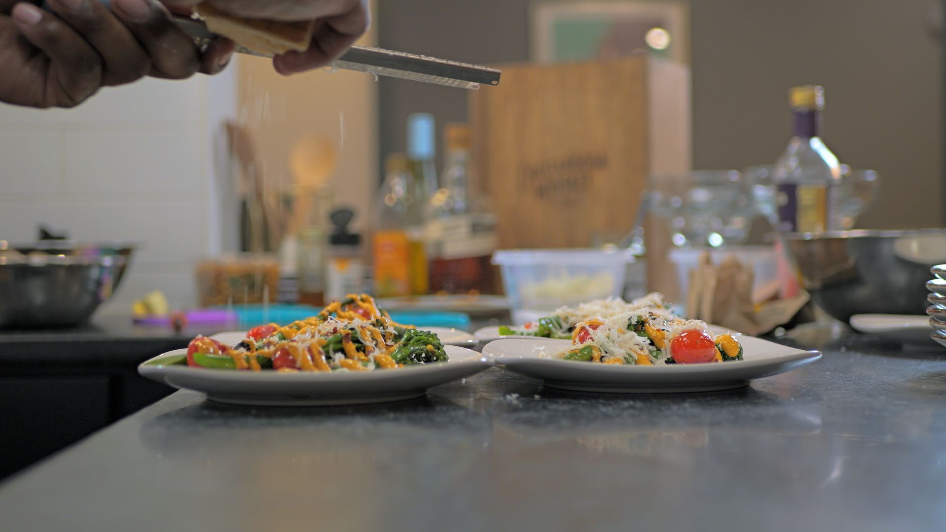 Two plated dishes on a kitchen counter, with a hand sprinkling herbs over one plate.