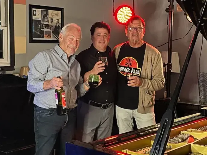 three men are posing for a picture in front of a piano . one of the men is wearing a jurassic park shirt