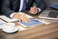 Person writing in a notebook beside a calculator and stacks of cash on a desk