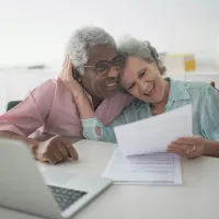 Two smiling older adults lean together over papers at a desk with a laptop nearby.