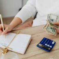 Person writing in a notebook beside a calculator and stacks of cash on a desk