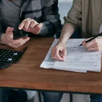 Two people reviewing documents at a wooden table, one using a phone and the other writing with a pen.