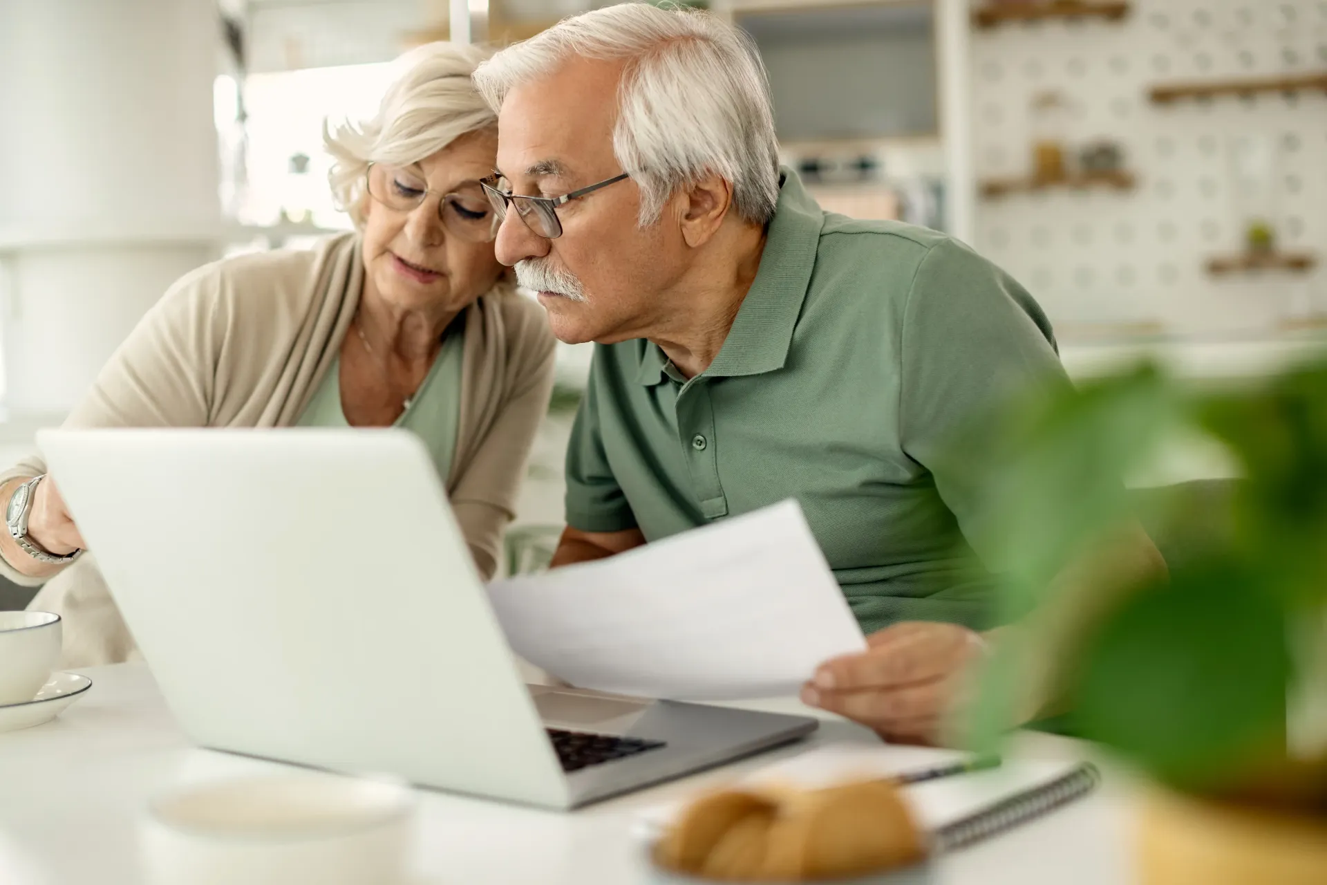 Older couple leaning over a laptop at a kitchen table, reviewing papers together