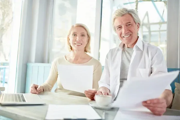 Two smiling people at a bright desk, reviewing documents with a laptop and coffee cups