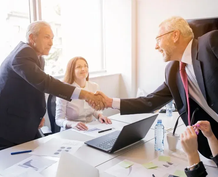 Business meeting with two people shaking hands across a conference table with a laptop and documents.
