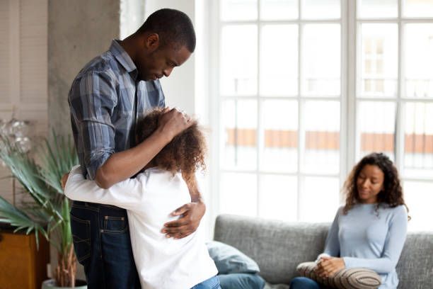 A man is hugging a little girl in a living room while a woman sits on a couch.