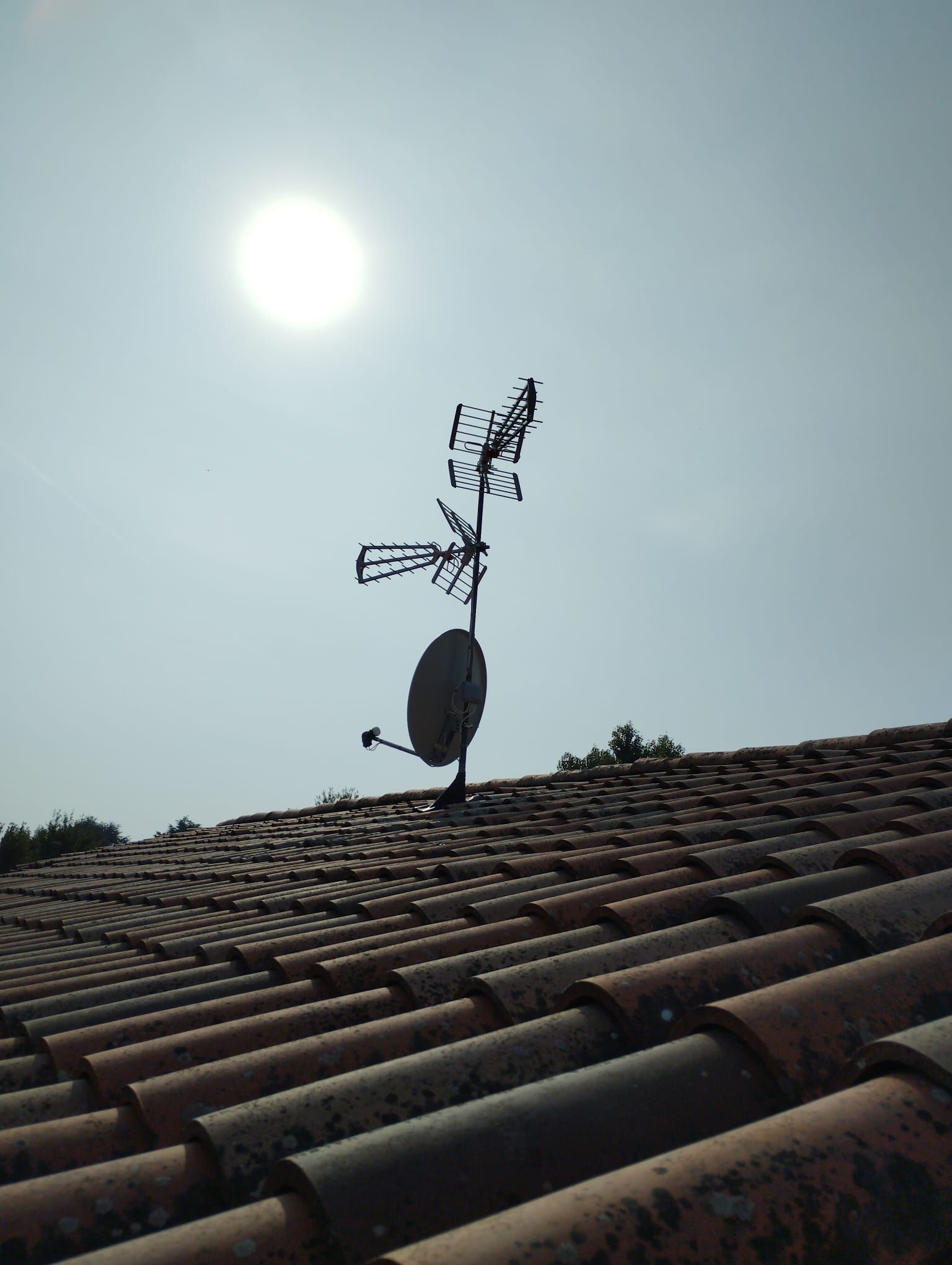 Antenna TV e parabola satellitare su un tetto di tegole di terracotta, sullo sfondo di un sole splendente e di un cielo azzurro.