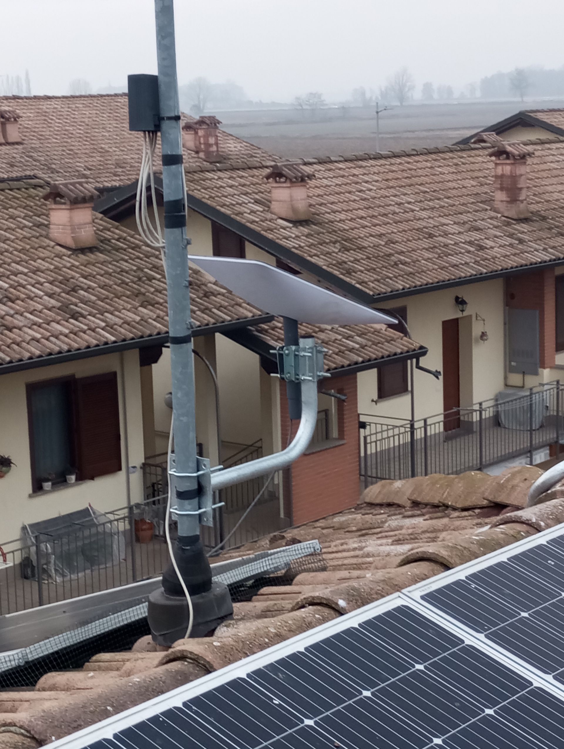 Antenna sul tetto con pannelli solari e case sullo sfondo in una giornata nuvolosa.