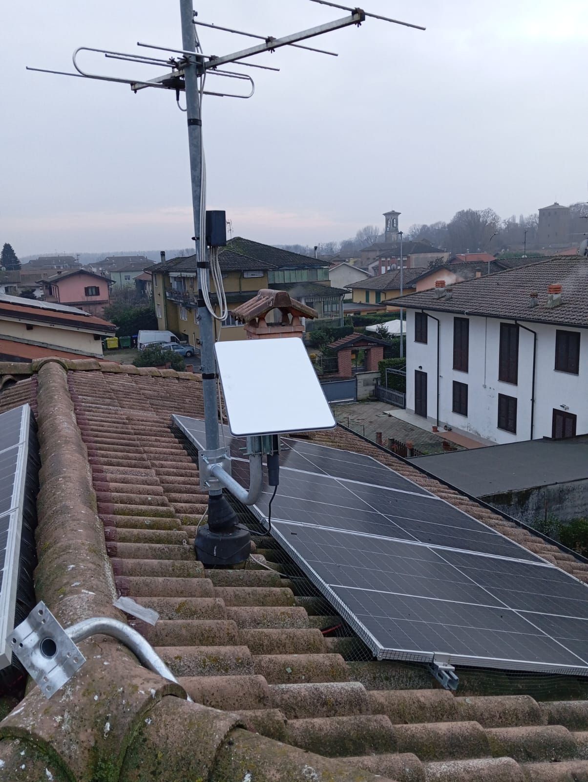 Tetto con pannelli solari, un'antenna e un dispositivo elettronico. Sullo sfondo, case e un cielo nuvoloso.