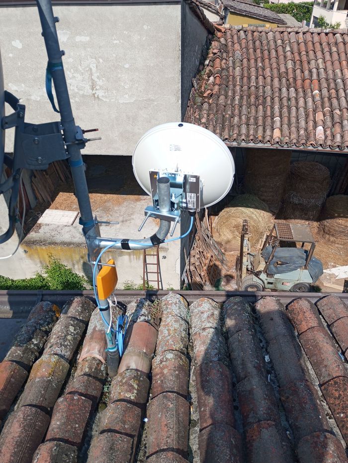 Un'antenna parabolica bianca montata sul tetto di un edificio, con un tetto di tegole e altre strutture visibili.