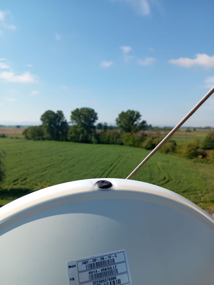 Vista dall'alto, su un campo verde con alberi e cielo azzurro; in primo piano c'è un'antenna parabolica.