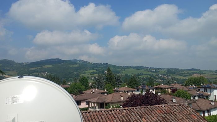 Vista di una città con tetti di tegole rosse e verdi colline sotto un cielo azzurro parzialmente nuvoloso.