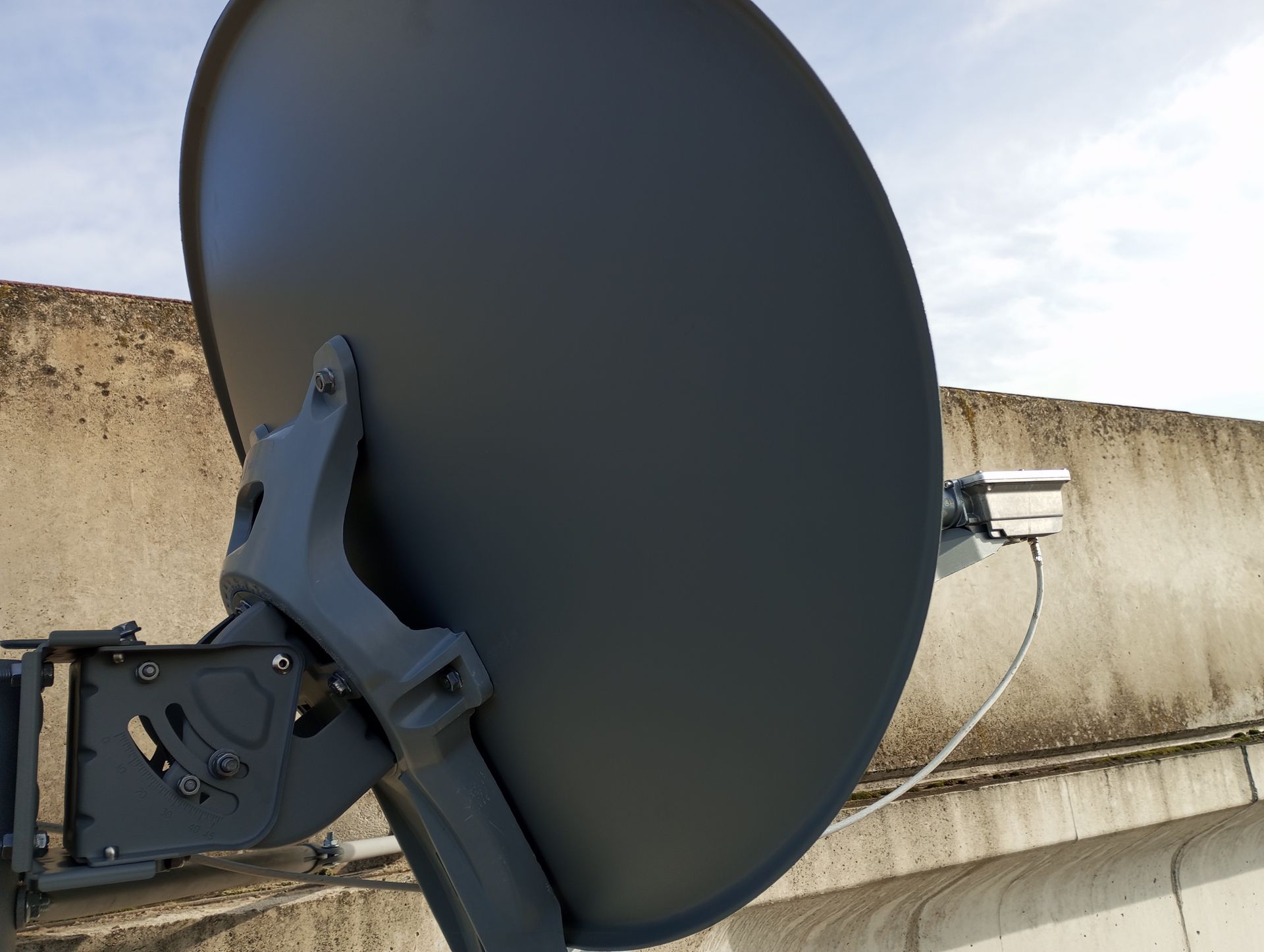 Parabola satellitare grigio scuro montata su un muro di cemento contro un cielo nuvoloso.