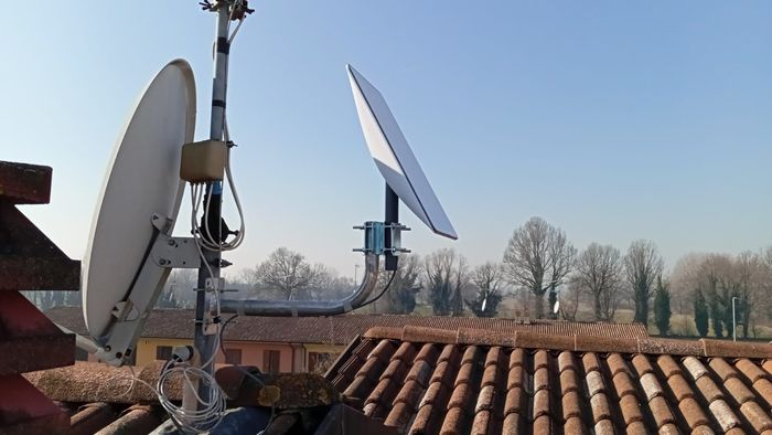 Due parabole satellitari su un tetto contro un cielo azzurro; una parabola rettangolare bianca è rivolta verso il cielo, mentre l'altra è una tipica parabola rotonda.