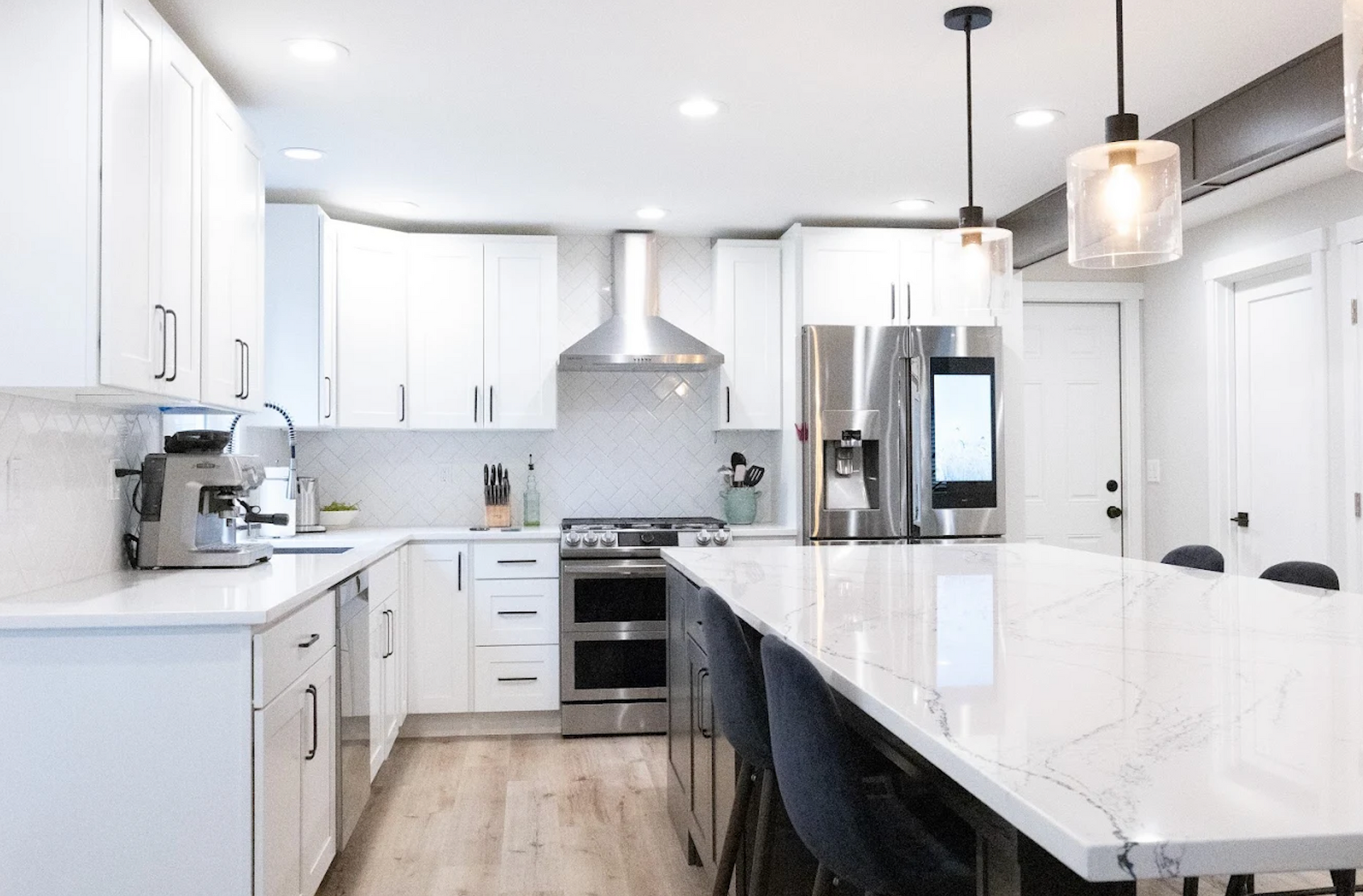Modern white kitchen with stainless steel appliances, marble countertops, and island with seating.