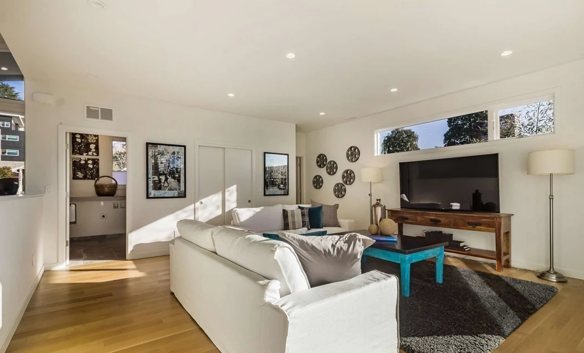 Bright living room with white sofa, blue table, and TV. Wooden floors, natural light.
