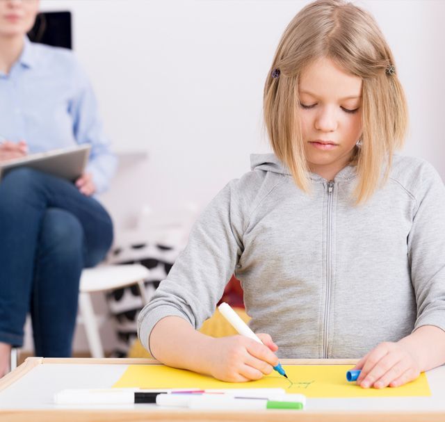 Una niña dibuja con un rotulador en una mesa; al fondo hay un terapeuta sentado.