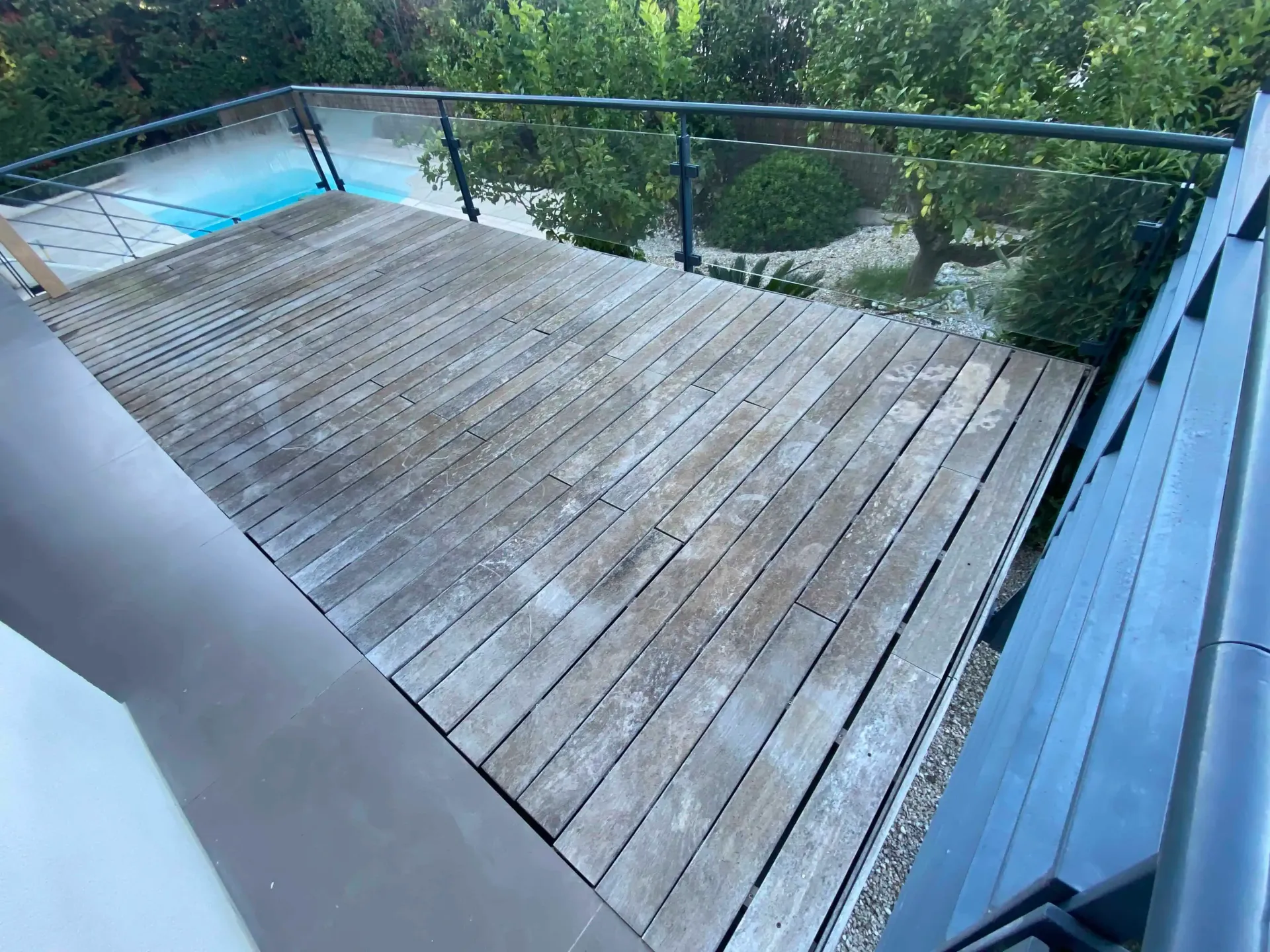Une terrasse tres usée en bois avec balustrade en verre donne sur une piscine et un jardin.