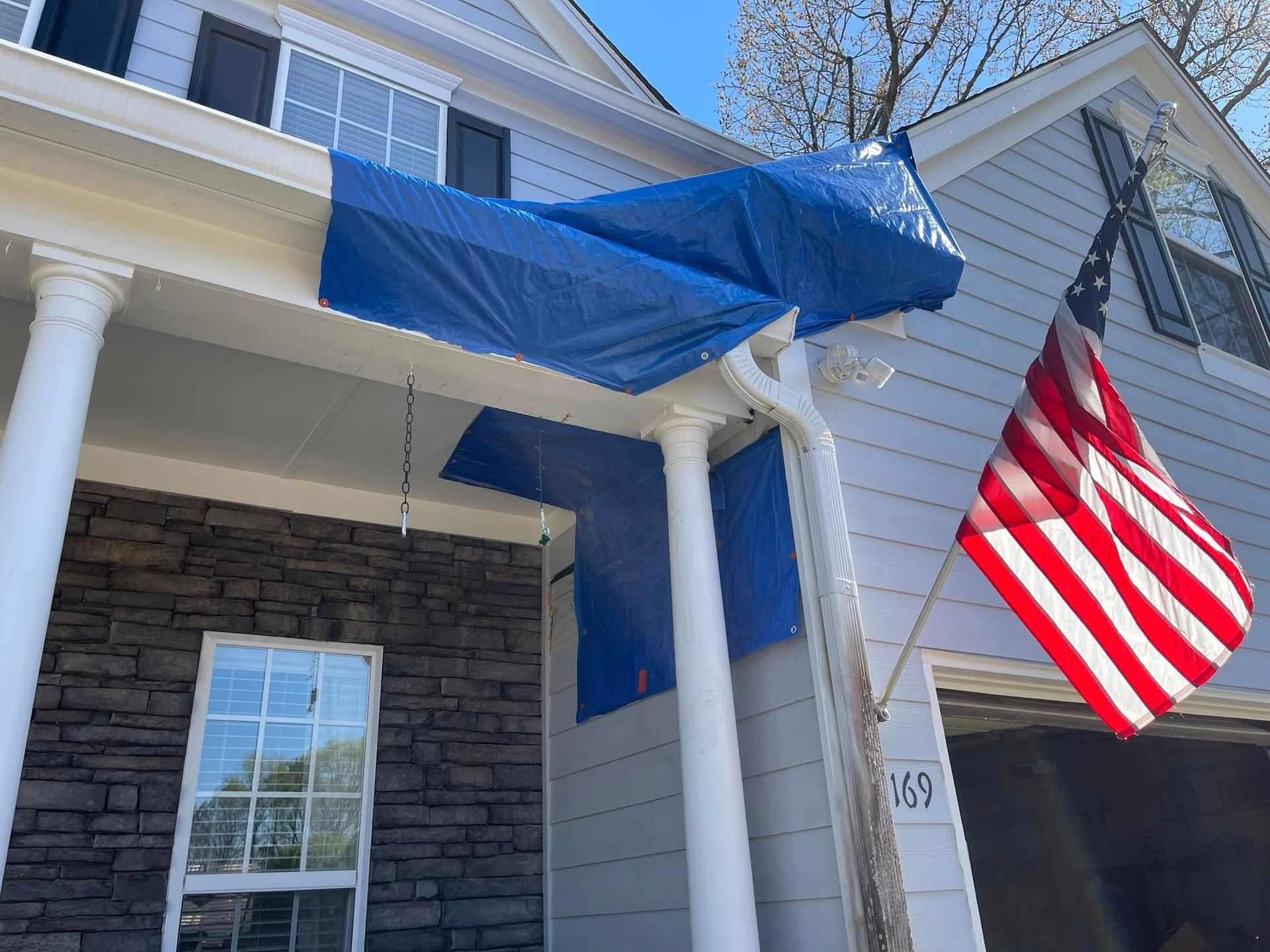 Blue tarp draped over a house porch, with a large American flag hanging beside white columns.