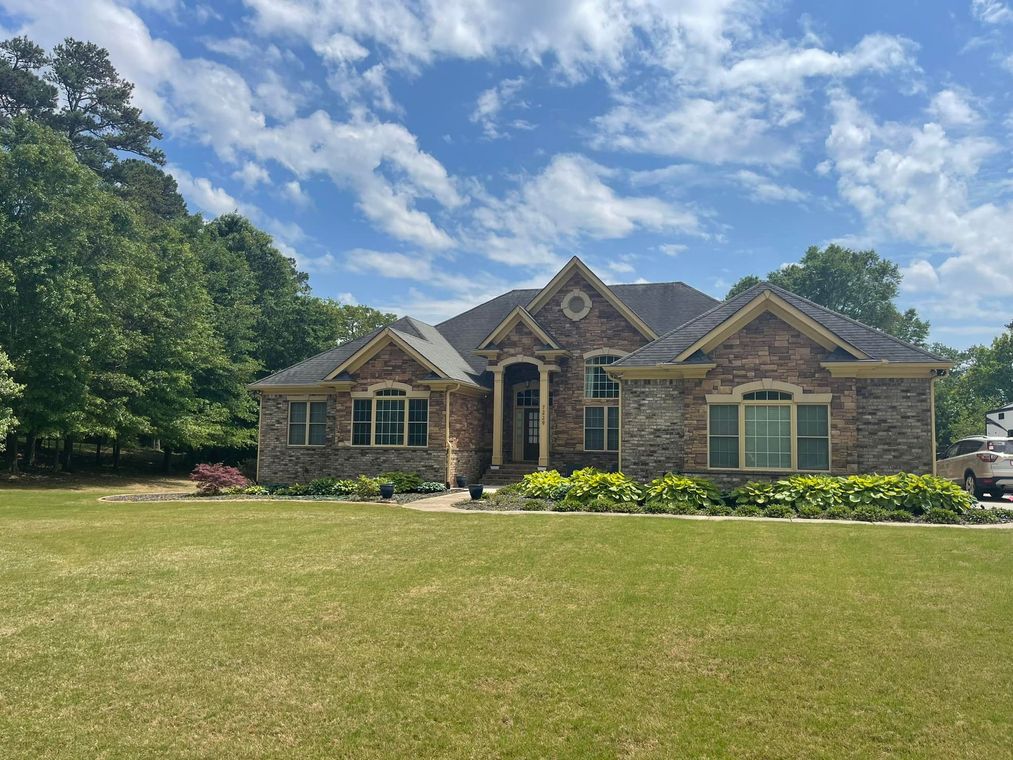 Brick suburban house with a wide lawn, trees, and a blue sky with scattered clouds