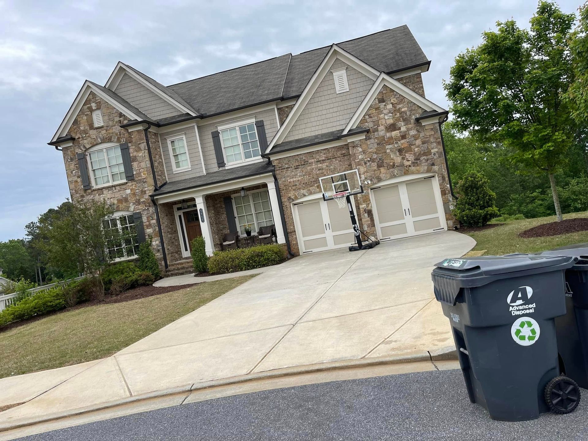 Large brick two-story suburban house with a wide driveway and a trash bin in the foreground.