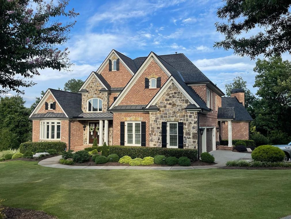 Large brick suburban house with dark roof, front lawn, and landscaped shrubs under a blue sky
