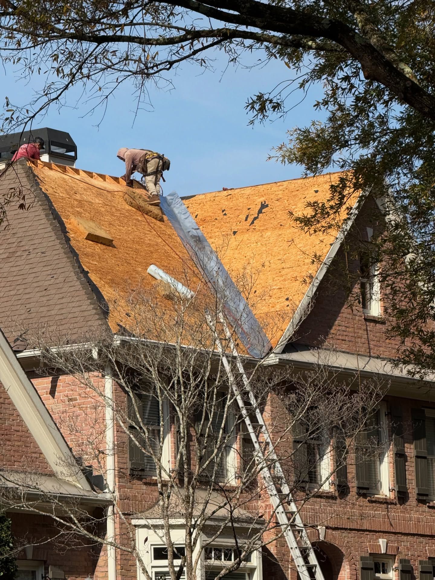 Workers on a rooftop replacing orange shingles with a ladder against a brick house