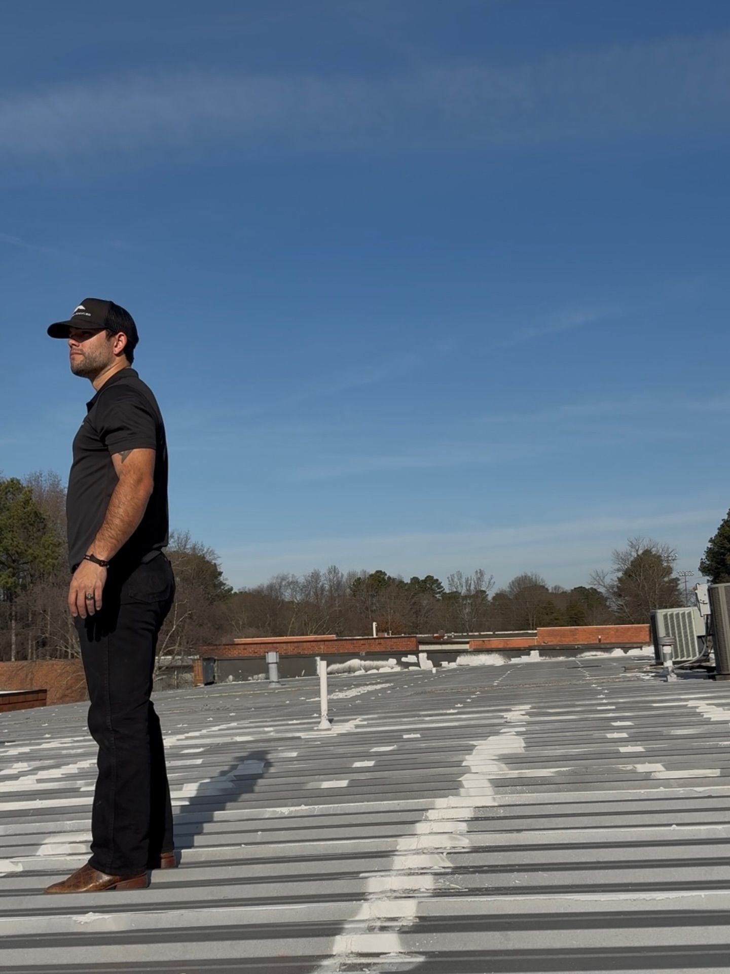 Person standing on a rooftop under a blue sky, wearing black clothing and a cap.