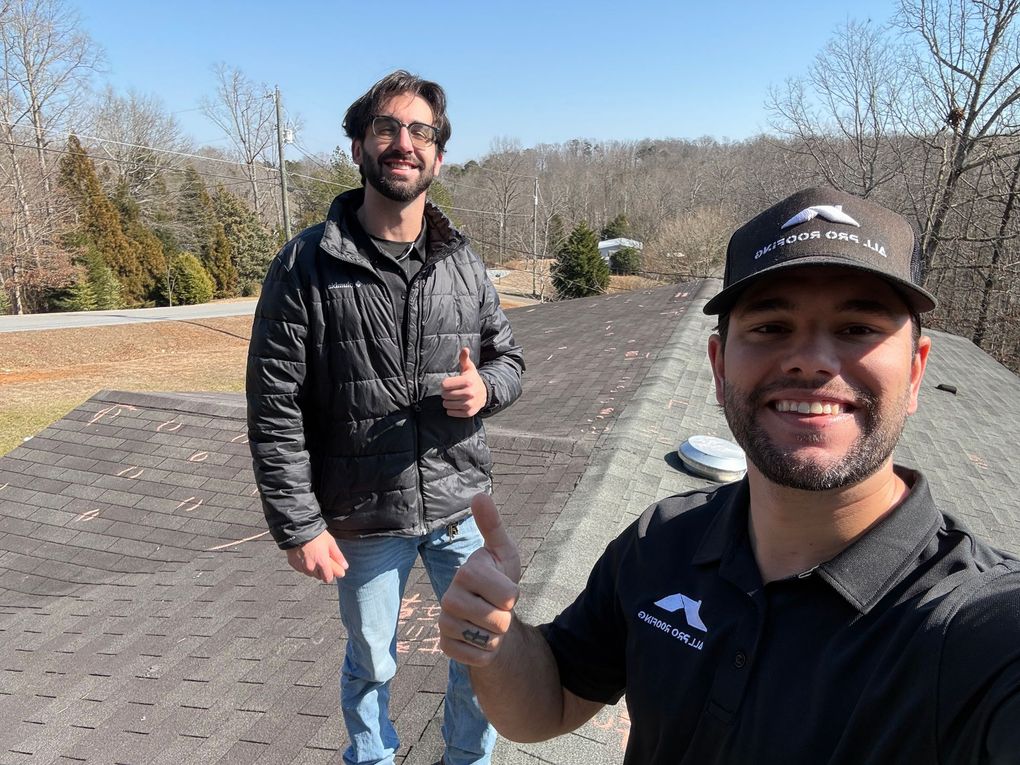Two men outdoors on a sunny path, smiling and giving thumbs up, with bare trees and parked cars behind them
