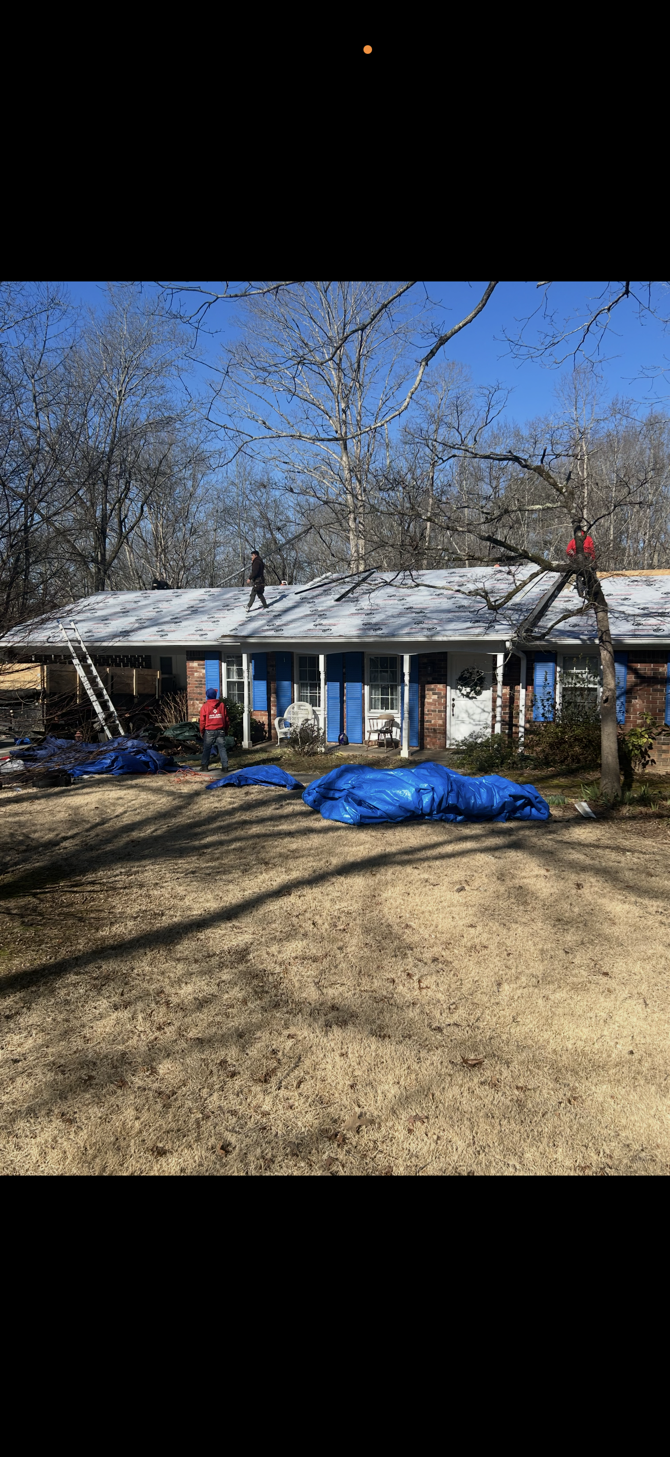 Single-story house with bare trees, blue tarp, and a person on the porch in a yard