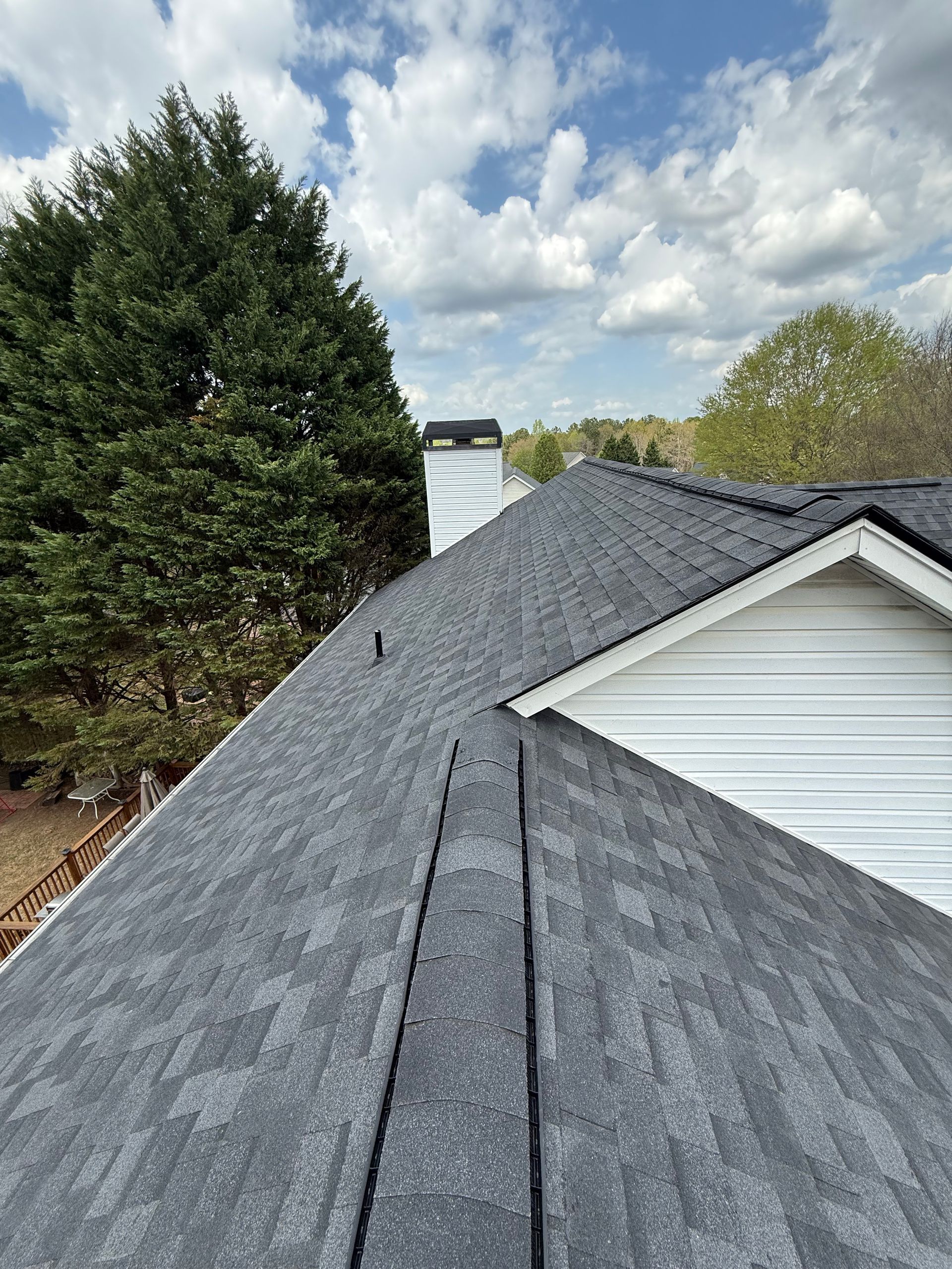 Gray shingled roof with a white house gable and tall trees under a cloudy sky