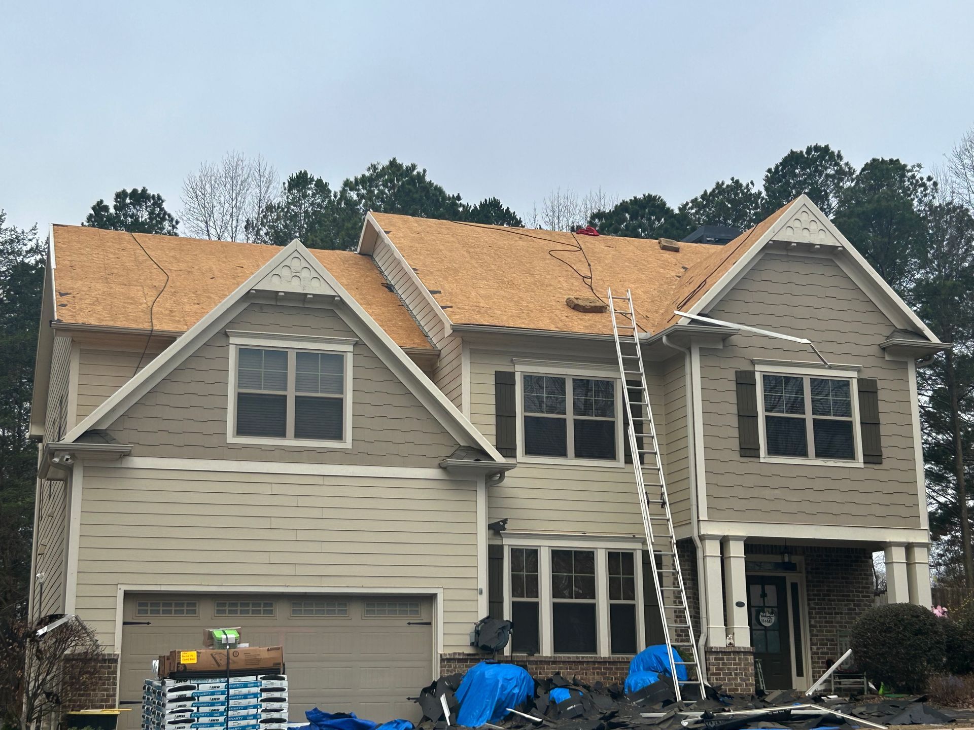 Two-story house under construction with exposed roof sheathing, ladders, and blue tarps in the front yard