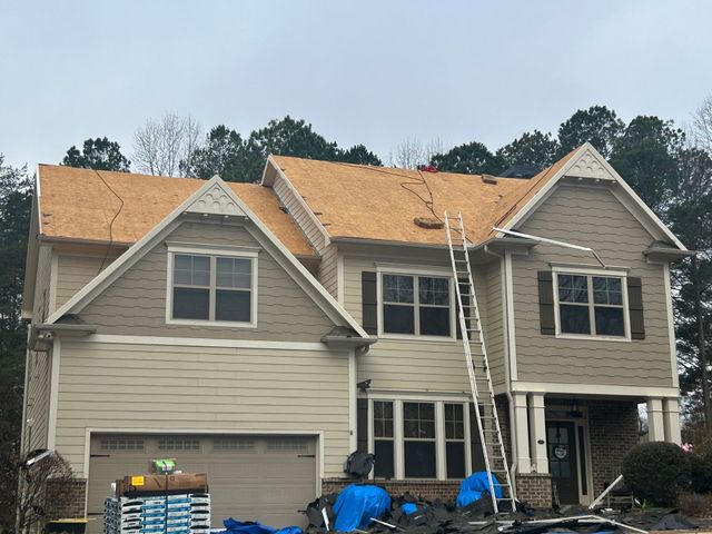 Two-story house under construction with exposed roof sheathing, ladders, and blue tarps in the front yard