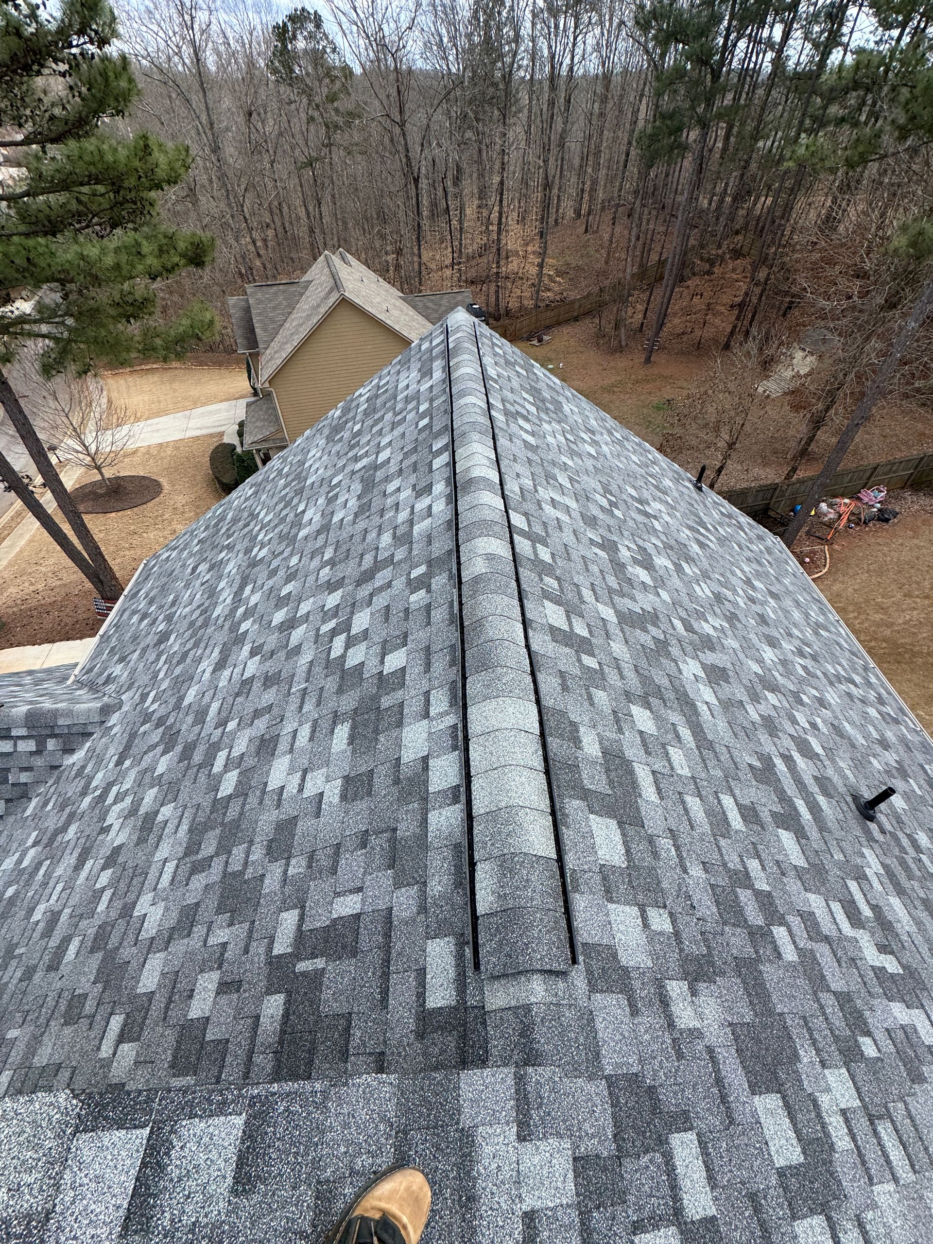Roof view with gray shingles and a tan outbuilding below, surrounded by leafless trees.