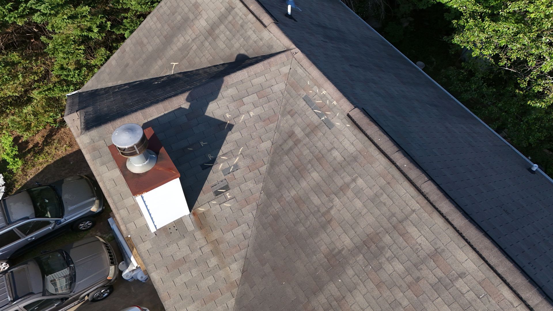 Aerial view of a shingled roof with a chimney and parked vehicles beside it.