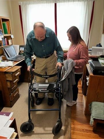 Man using a walker, assisted by a woman, indoors, near a desk.