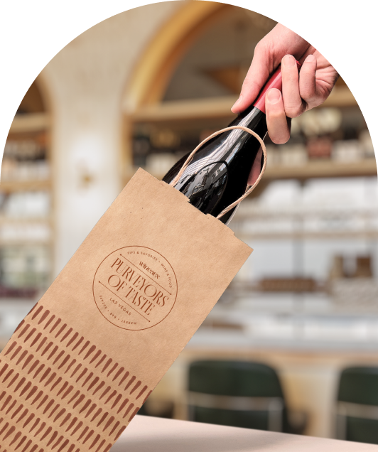 A hand pulls a wine bottle from a brown paper shopping bag labeled