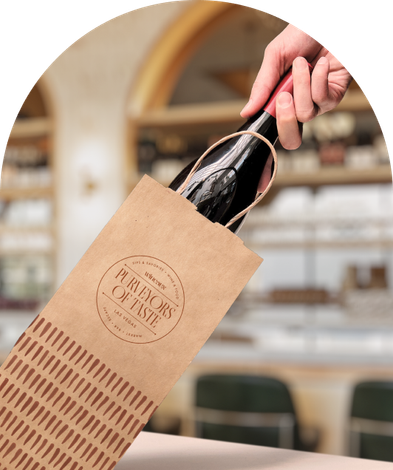 A hand pulls a wine bottle from a brown paper shopping bag labeled